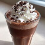 Close-up of a glass filled with thick, frosty frozen hot chocolate topped with whipped cream and chocolate shavings.