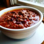 Close-up of a steaming white bowl filled with rich, meaty chili containing kidney beans.
