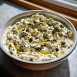 A creamy, chunky dirty martini dip topped with olives, herbs, and olive oil in a white bowl.