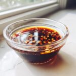 A small glass bowl filled with dark, savory Dumpling Sauce topped with chili flakes and garlic bits.