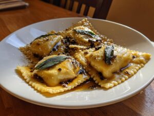 Four large, square beef short rib ravioli served on a white plate with brown butter sauce, grated cheese, and sage leaves.