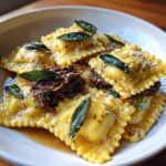 Close-up of homemade beef short rib ravioli tossed in brown butter sauce, topped with shredded meat, sage leaves, and Parmesan.