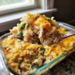 A spoonful of creamy chicken and rice casserole topped with melted cheddar cheese, lifted above the baking dish.