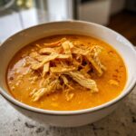 A close-up of a bowl of rich, orange-colored chicken tortilla soup topped with shredded chicken and crispy tortilla strips.