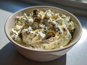 A close-up of a creamy, savory dirty martini dip topped with sliced green olives and herbs.