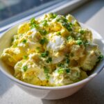 Close-up of creamy egg salad in a white bowl, garnished generously with chopped green chives.