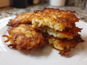 A close-up of golden brown, crispy potato latkes stacked on a white plate, one broken open to show the shredded interior.