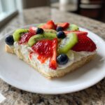 A square slice of fruit pizza sugar cookie topped with whipped cream, strawberries, kiwi, and blueberries.