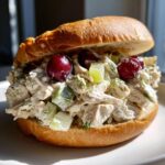 Close-up of a gourmet chicken salad sandwich overflowing with creamy filling, celery, grapes, and dill on a crusty roll.