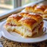 A serving of four baked Ham and Cheese Sliders with gooey, melted cheese stretching out from the golden brown rolls.