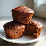 Three dark, moist bran muffins stacked on a white plate, one is broken open showing the texture.
