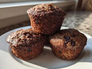 Three dark, moist bran muffins, studded with dried fruit, stacked on a white plate in bright sunlight.