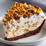 A thick slice of no-bake butterfinger pie featuring a chocolate crust, creamy filling with candy pieces, and a whipped topping.