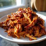 Close-up of saucy, shredded pioneer woman slow cooker pulled pork piled high on a white plate.