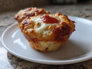 Two golden-brown pizza muffins topped with melted, bubbly cheese and a dollop of sauce, served on a small white plate.