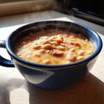 Close-up of a steaming blue mug filled with creamy white bean and ham soup, highlighted by sunlight.