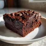 A close-up, rich, dark slice of sweet potato brownies topped with chocolate chips, sitting on a white plate.