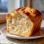 A thick slice of golden-crusted beer bread showing its soft, fluffy interior, resting on a white plate.
