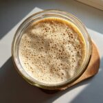 Overhead view of an active sourdough starter in a glass jar, showing many small, uniform bubbles indicating readiness.