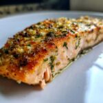 Close-up of a perfectly cooked, flaky air fryer salmon fillet topped with garlic and herbs on a white plate.