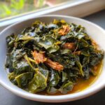 Close-up of tender, braised collard greens served in a white bowl with savory pot liquor and pieces of smoked meat.