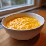 Close-up of a bowl filled with bright orange, melted beer cheese dip resting on a wooden cutting board near a window.