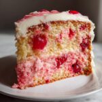 Close-up of a moist slice of cherry cake featuring yellow sponge layers, pink cherry filling, and white frosting.