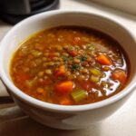 Close-up of a white bowl filled with rich, savory lentil soup featuring carrots, celery, and herbs.