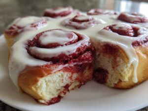A close-up of several moist red velvet cinnamon rolls covered in thick white cream cheese icing.