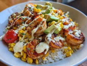 A close-up, vibrant photo of a delicious shrimp bowl featuring grilled shrimp, corn, avocado slices, and a creamy white sauce over rice.