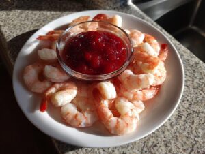 A white plate featuring cooked shrimp arranged around a small glass bowl of bright red shrimp cocktail sauce.