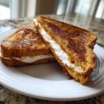 Two halves of perfectly golden brown stuffed french toast filled with creamy white filling on a white plate.