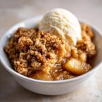 Close-up of a warm serving of apple crisp recipe topped with a scoop of vanilla ice cream in a white bowl.