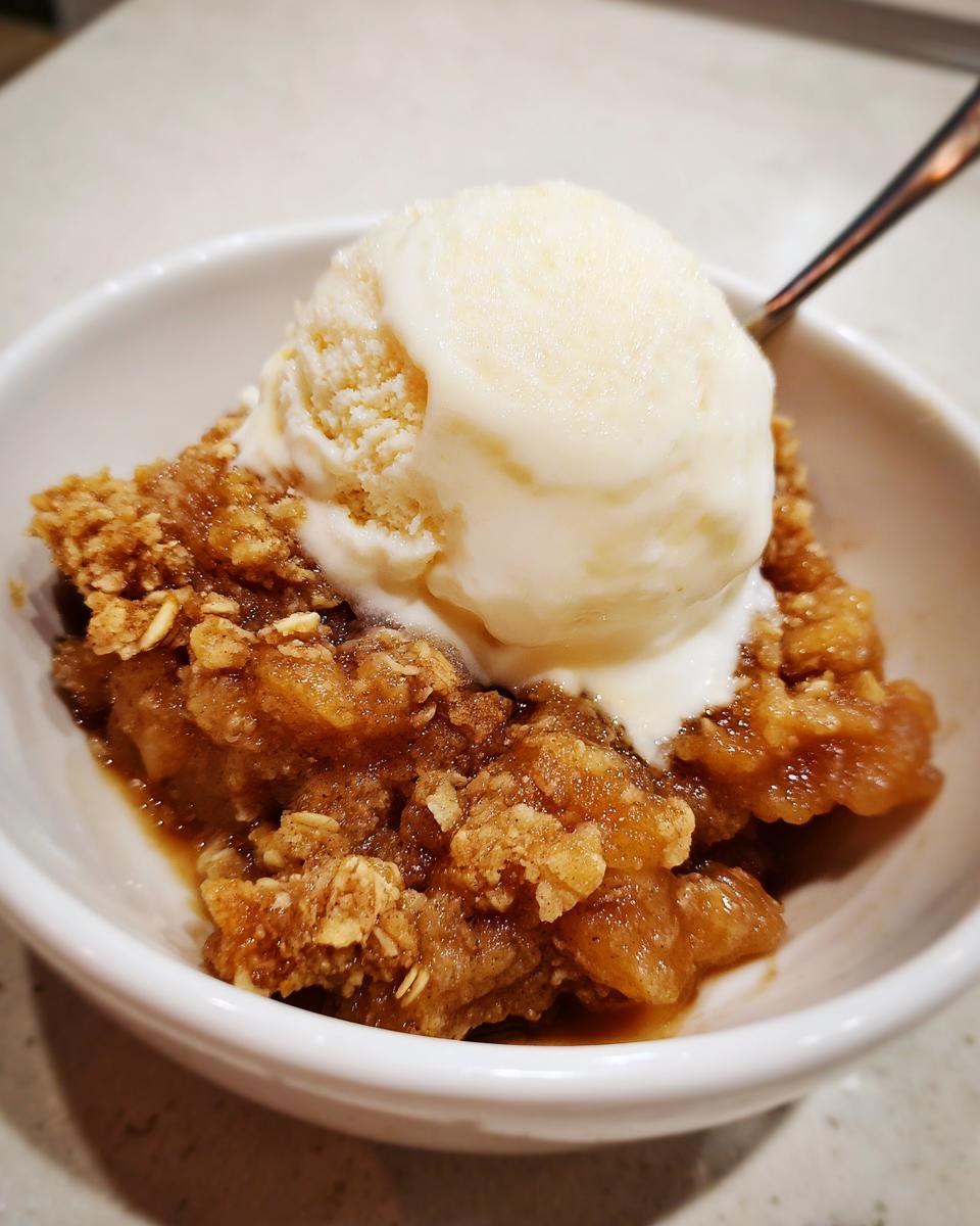 Close-up of a warm serving of apple crisp recipe topped with a melting scoop of vanilla ice cream in a white bowl.
