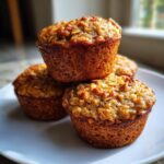 A stack of three golden-brown banana oatmeal muffins topped with oats, sitting on a white plate.
