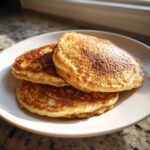 Three golden-brown banana oatmeal pancakes stacked on a white plate, catching bright sunlight.