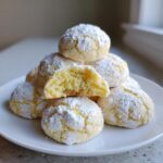 Stack of soft, crinkle-style lemon cookies heavily dusted with powdered sugar, one cookie is broken open.