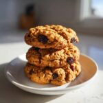 Three chewy brown butter oatmeal raisin cookies stacked on a small white plate, brightly lit by sunlight.