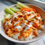 Close-up of a white bowl filled with white rice topped with saucy buffalo chicken, drizzled with ranch, and garnished with celery and carrots.