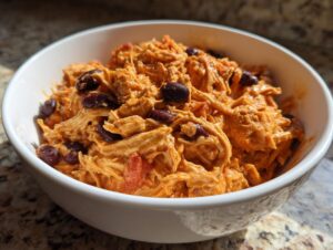 A close-up of a white bowl filled with creamy, shredded buffalo chicken chili mixed with dark red kidney beans.