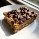 A single, thick, no-bake protein bars square topped generously with dark chocolate chips on a white plate.