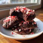 A stack of three rich, dark chocolate peppermint cookies topped with crushed candy canes on a white plate.