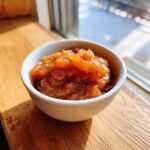 A small white bowl filled with chunky, spiced applesauce, sitting on a wooden surface near a bright window.