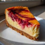 A close-up of a slice of cranberry orange cheesecake with a graham cracker crust and bright red fruit swirl.