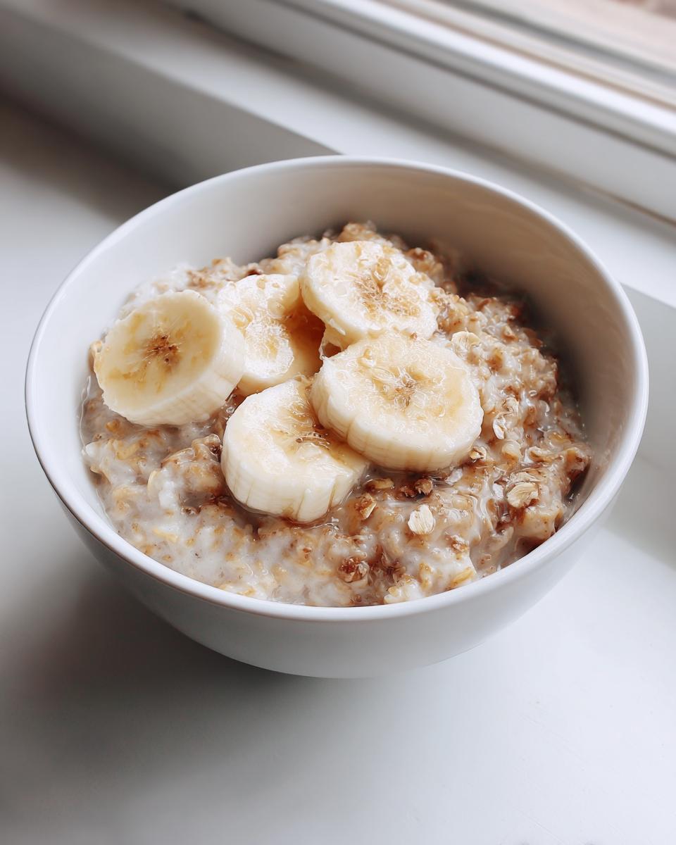 A white bowl filled with creamy banana oatmeal topped with fresh banana slices and a drizzle of syrup.