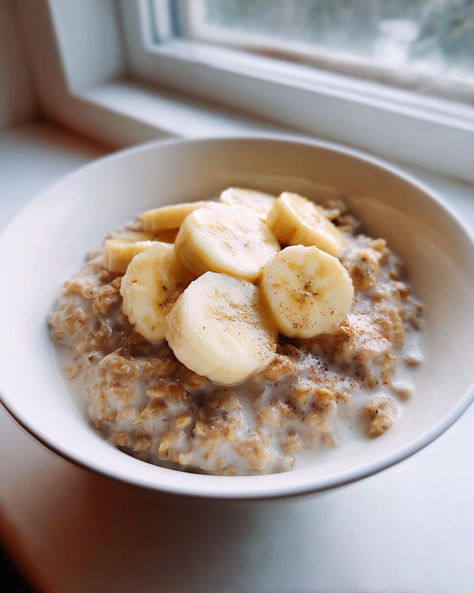 A close-up of creamy banana oatmeal in a white bowl, topped with fresh banana slices and a sprinkle of cinnamon.