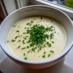 A close-up of a bowl of smooth, creamy cauliflower soup garnished generously with chopped fresh chives and black pepper.