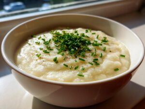A close-up of creamy cauliflower soup in a white bowl, garnished with fresh chopped chives and black pepper.