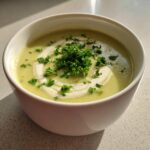 A bowl of creamy, light green celery soup garnished with a swirl of cream and fresh chopped parsley.