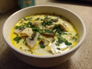 Close-up of a creamy, yellow-tinged coconut soup featuring chunks of chicken, sliced mushrooms, and fresh cilantro.